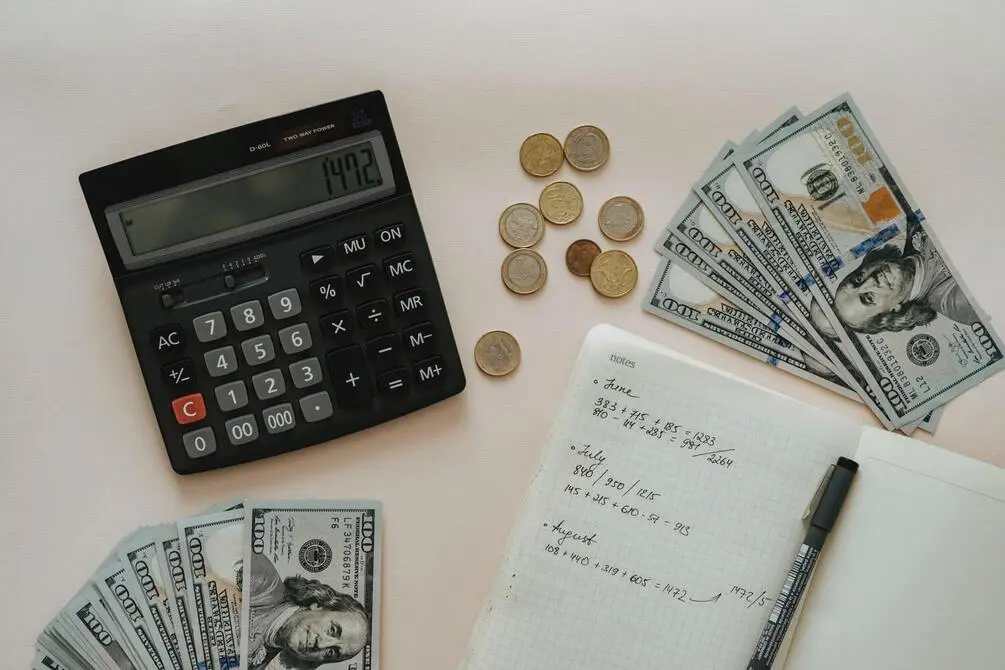 Desk with calculator, notepad, pen and money laid out on it.